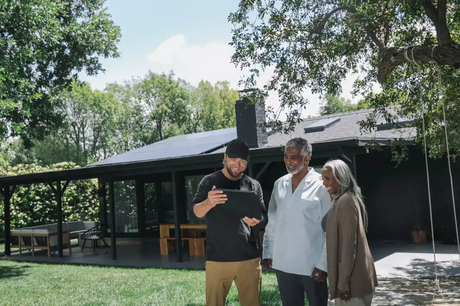 A solar installer reviewing system details on a tablet with two homeowners outside their solar-equipped home