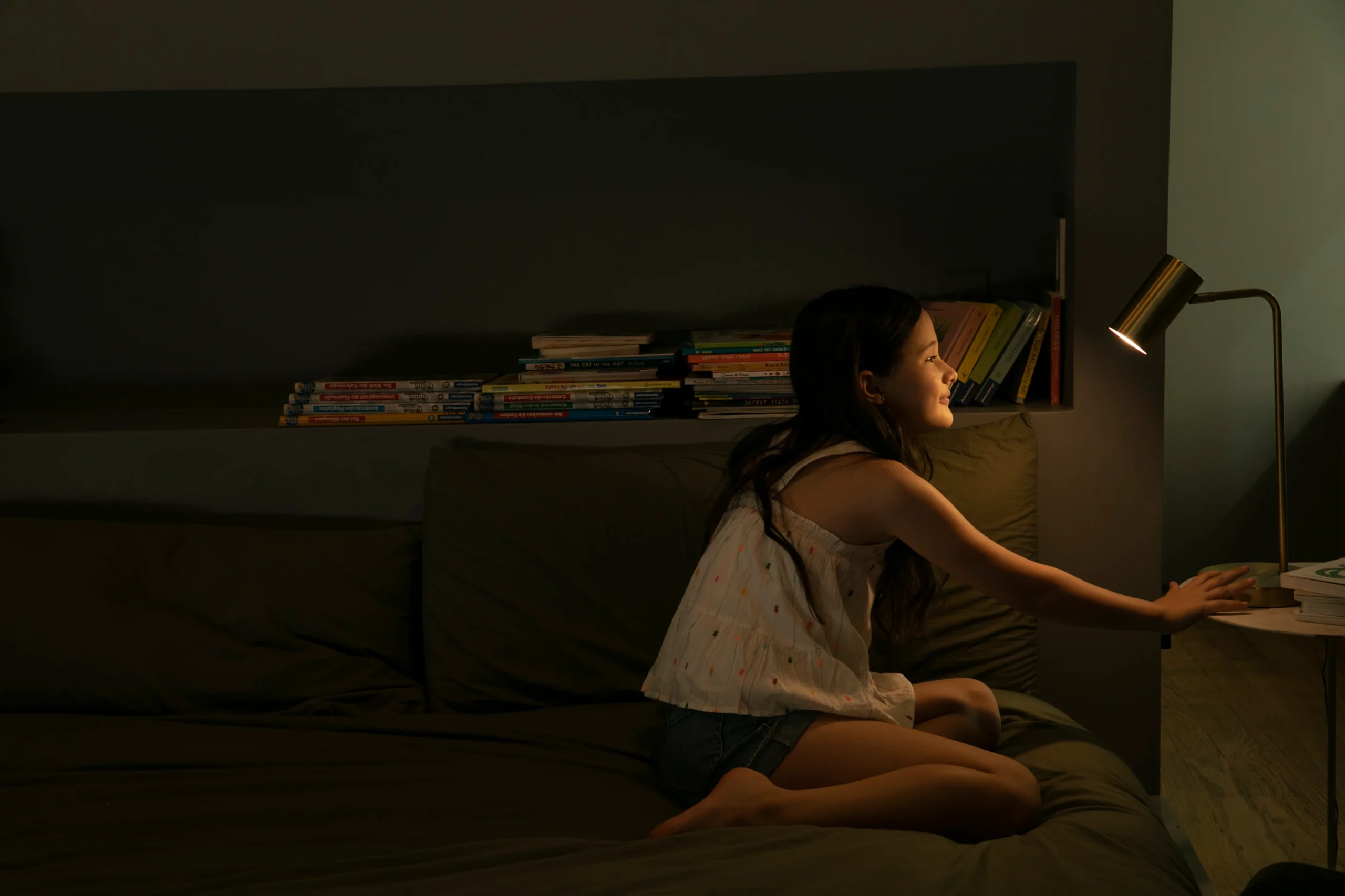 A young girl turns on a lamp in a dark room that sits in front of her bookshelf.