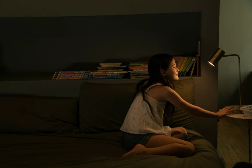 A young girl turns on a lamp in a dark room that sits in front of her bookshelf.