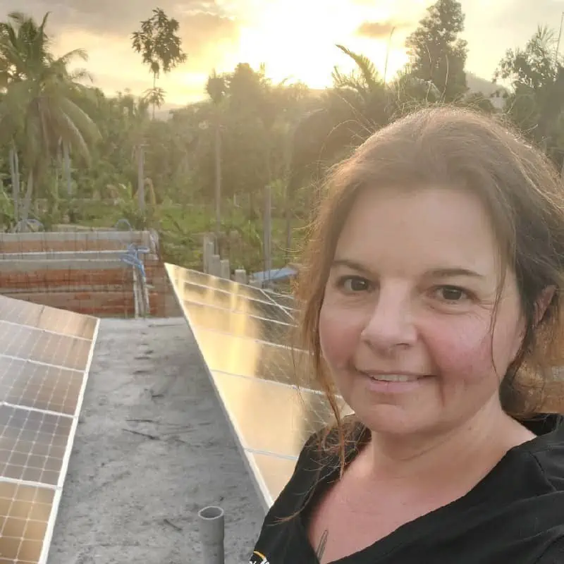 A woman with a content smile standing in front of solar panels with a tropical backdrop basking in the golden hour sunlight.